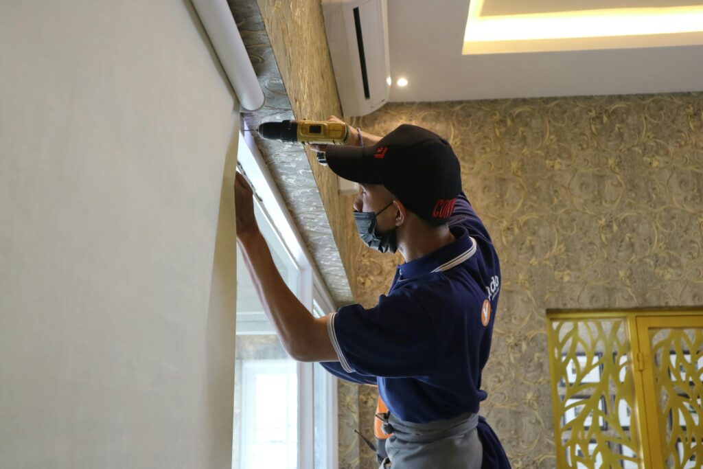 A worker using a drill to install window blinds in an indoor setting.